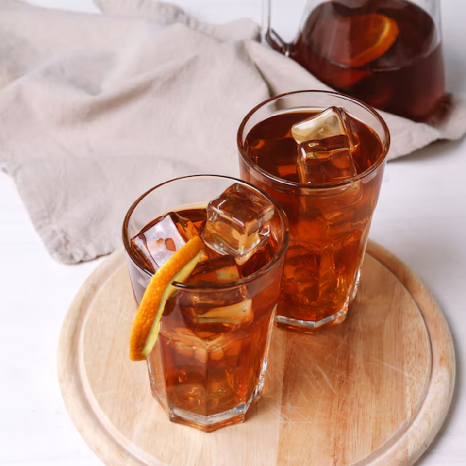 Two glasses of iced tea with orange slices and a pitcher on a wooden board.