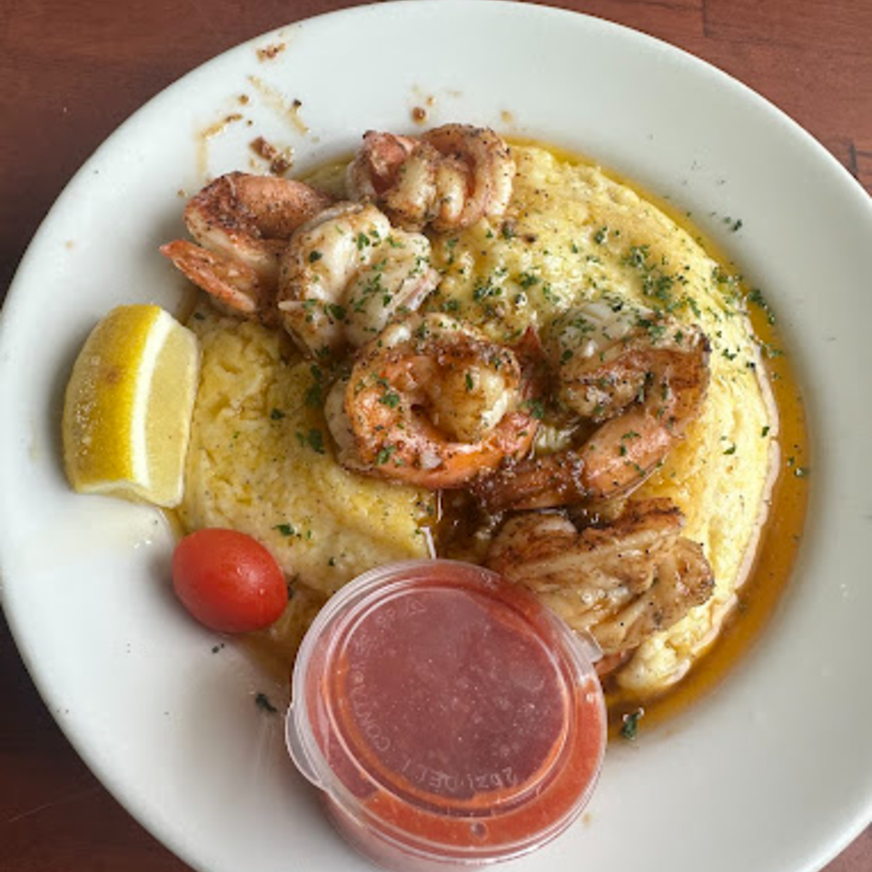 Shrimp and grits with a lemon wedge, cherry tomato, and a side of sauce on a white plate.